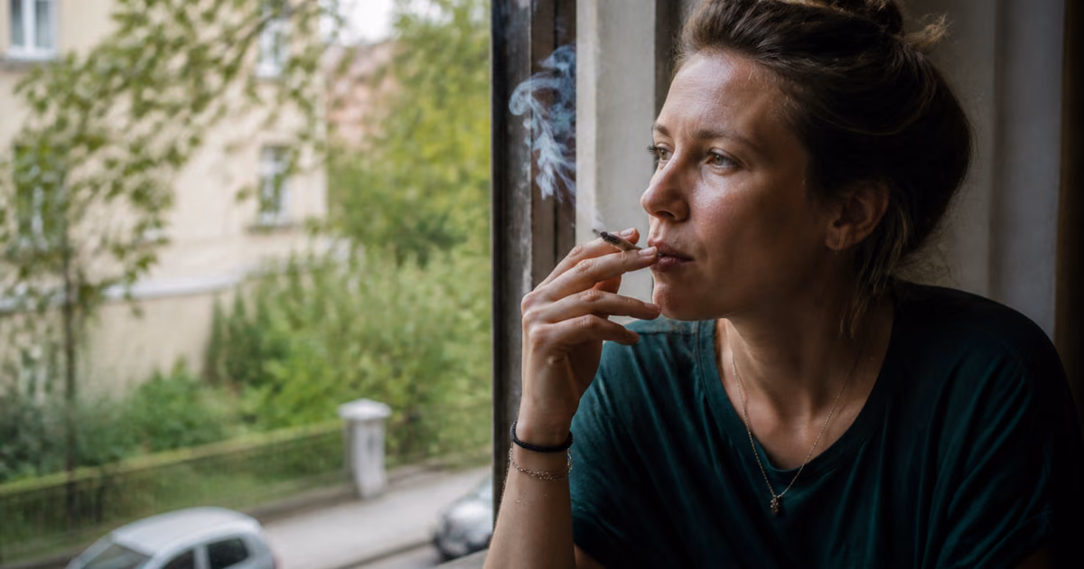 Kobieta pali jointa przy oknie, patrząc na zieloną ulicę